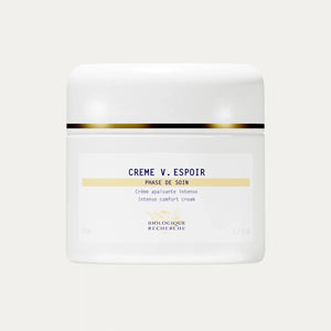 A white jar of Crème V. Espoir with gold detailing, described as an intense comfort cream formulated to calm reactive skin and reduce redness for a more even complexion.
