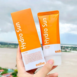 Close-up of a hand holding an orange tube and box of "Esthemax Hydrosun" sunscreen, set against a cloudy sky and river backdrop.