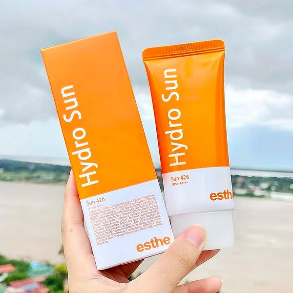 Close-up of a hand holding an orange tube and box of "Esthemax Hydrosun" sunscreen, set against a cloudy sky and river backdrop.