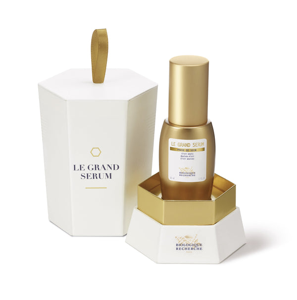 The Biologique Recherche Le Grand Serum - 1 oz, a gold bottle enriched with antioxidants for anti-aging benefits, is showcased next to its white and gold hexagonal packaging.