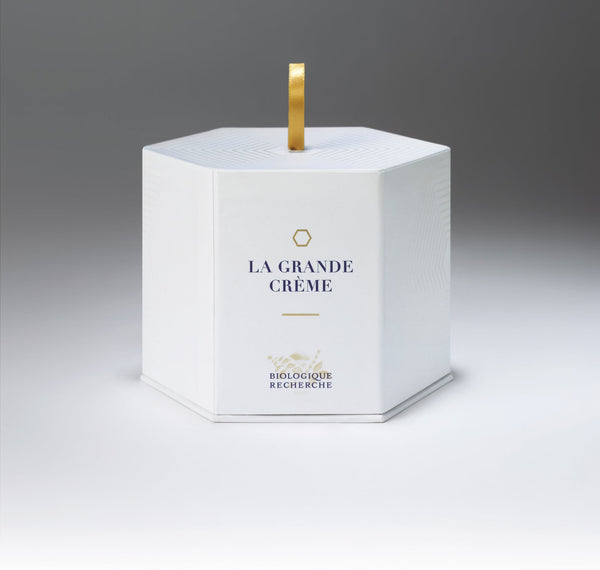 The hexagonal white box elegantly displays "Biologique Recherche La Grande Crème" and features a gold hexagon with a ribbon-like handle. This professional cosmetic promotes cellular regeneration and anti-aging effects.